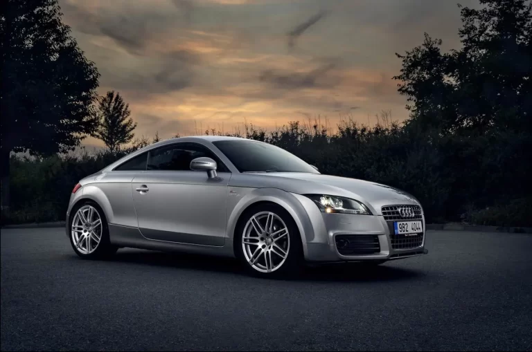 A silver Audi TT coupe is parked on a paved area with a dramatic sky and trees in the background.