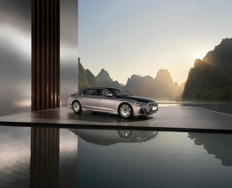 A silver luxury sedan is parked on a modern platform with a reflective water surface, set against a mountainous landscape at sunset.