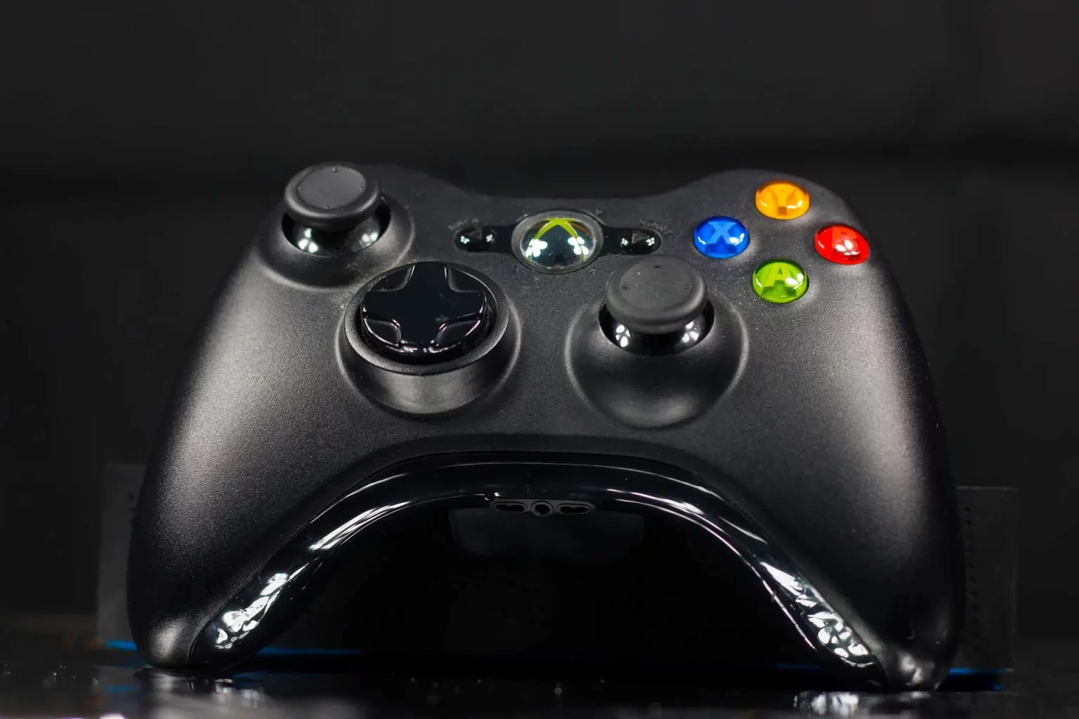 A close-up of a black gaming controller with colored buttons, two joysticks, and a directional pad, placed on a reflective surface against a dark background.