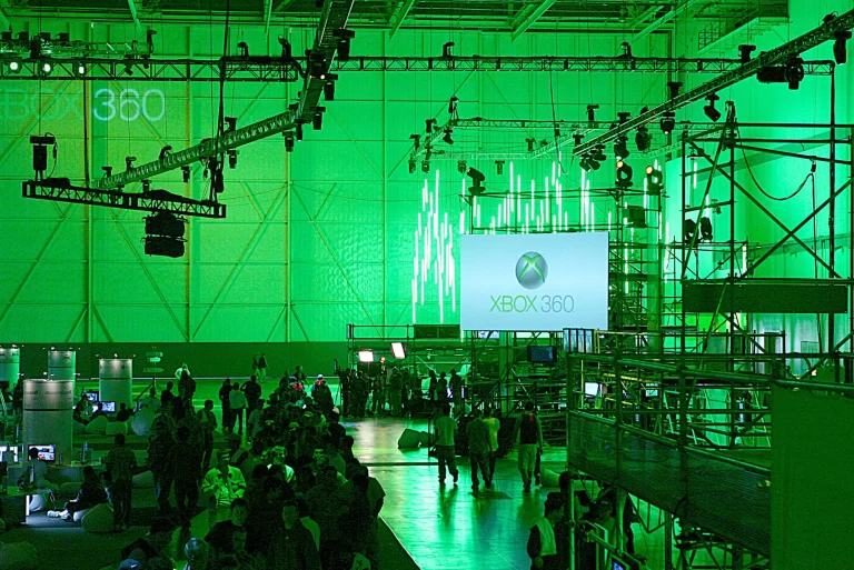 A crowded event space with green lighting, large Xbox 360 logos projected on walls, a central screen, and scaffolding structures around.
