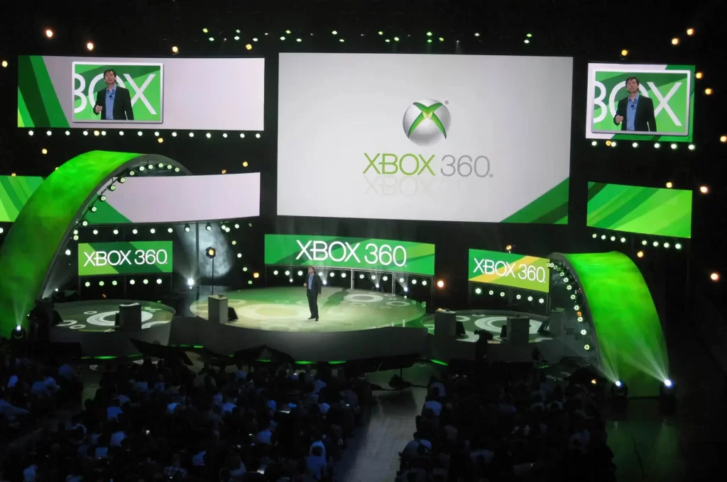 A person stands on a stage surrounded by multiple screens and lights displaying the Xbox 360 logo in front of an audience.