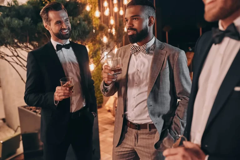 Three men in formal attire, one in a grey suit and two in black tuxedos, stand outdoors at night, holding drinks and conversing under string lights.