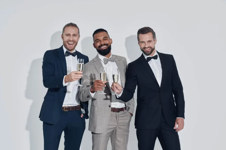 Three men in formal attire, one in a gray suit and two in black suits, stand side by side, holding champagne glasses and smiling against a plain white background.