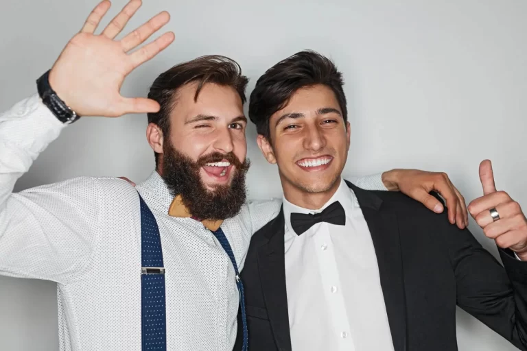 Two men in formal attire smiling; one man with a beard and wearing suspenders waves with one hand, while the other man gives a thumbs up.
