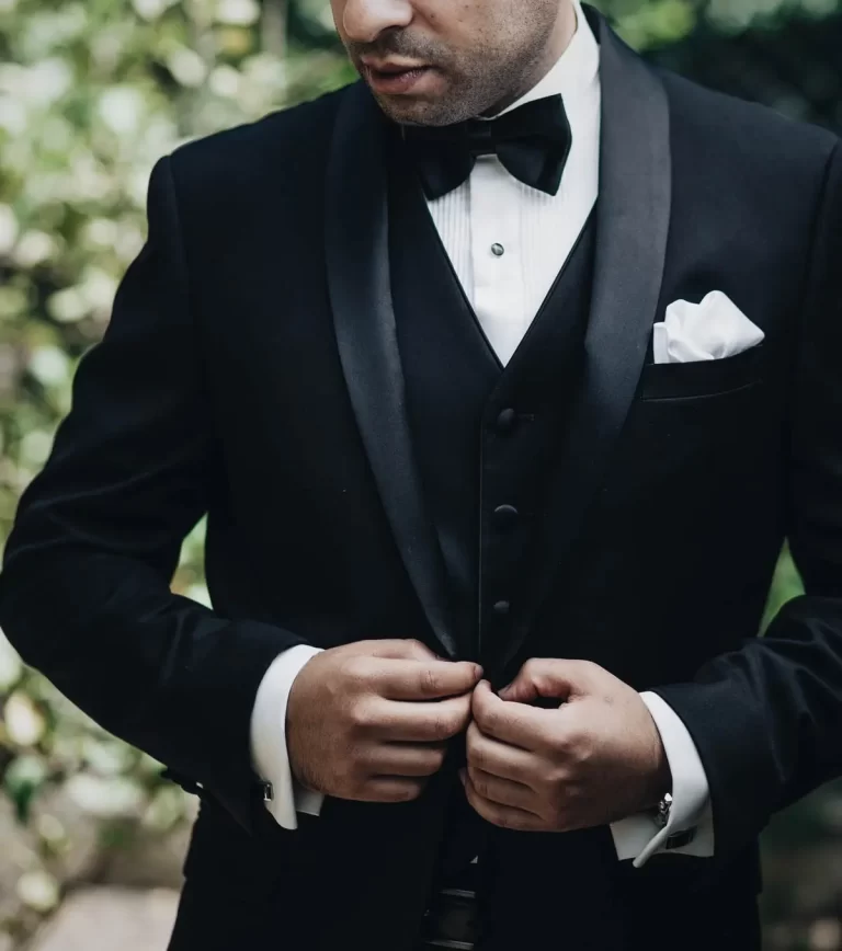 A man wearing a black tuxedo and bow tie adjusts his jacket outdoors.