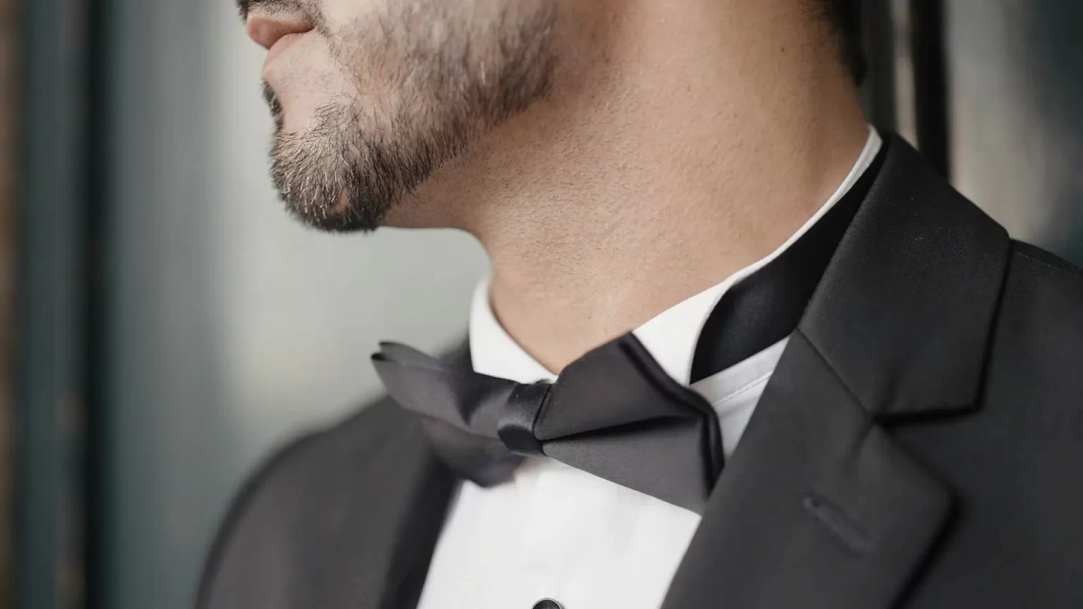 Close-up of a person wearing a black tuxedo, white dress shirt, and black bow tie. The shot focuses on the lower face and neck, showing a neatly trimmed beard.