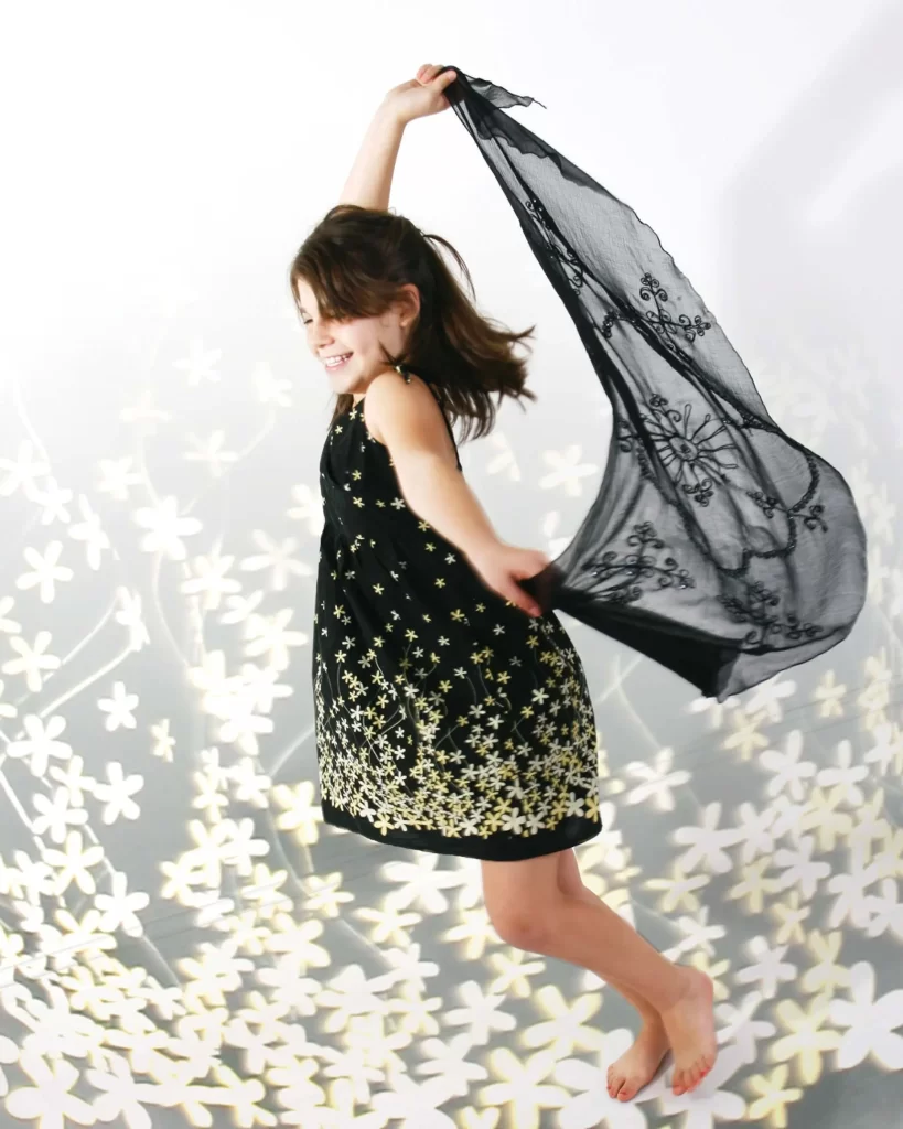 A young girl in a black dress with star patterns joyfully jumps, holding a black lace scarf. The background features a floral design with white flowers.