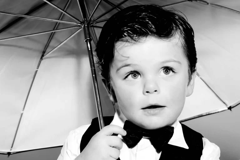 A young child dressed in a formal outfit holds an umbrella. The child has wide eyes and a curious expression. The image is in black and white.