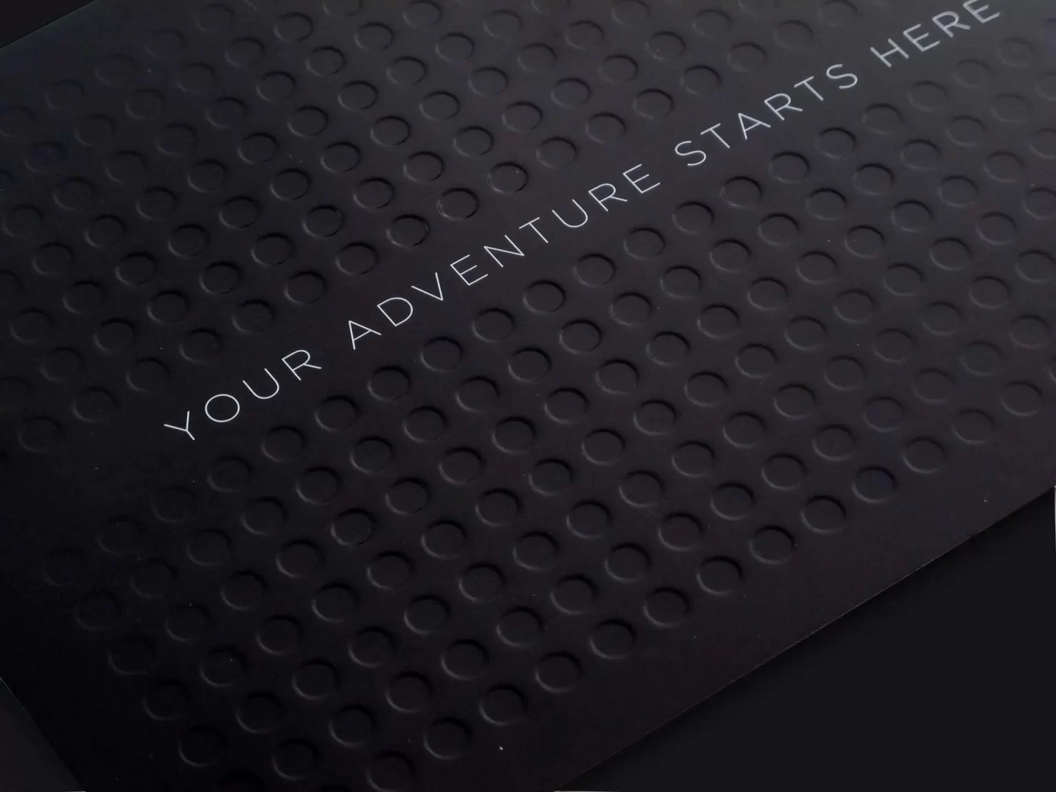 Close-up of a black textured surface with embossed circular patterns and the text "YOUR ADVENTURE STARTS HERE" printed in white.