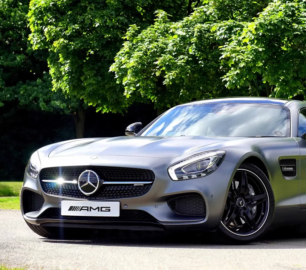 A silver Mercedes-Benz AMG GT sports car parked on a sunlit road with lush green trees in the background.