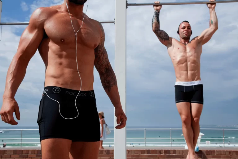 Two muscular men in black athletic shorts exercise outdoors; one is doing a pull-up, and the other stands listening to headphones. The ocean and sky are in the background.