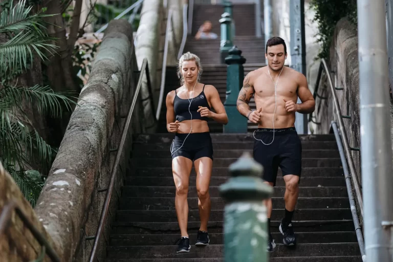 Two people jog down outdoor stairs while wearing black athletic wear and earphones. Trees and foliage border the stone staircase.