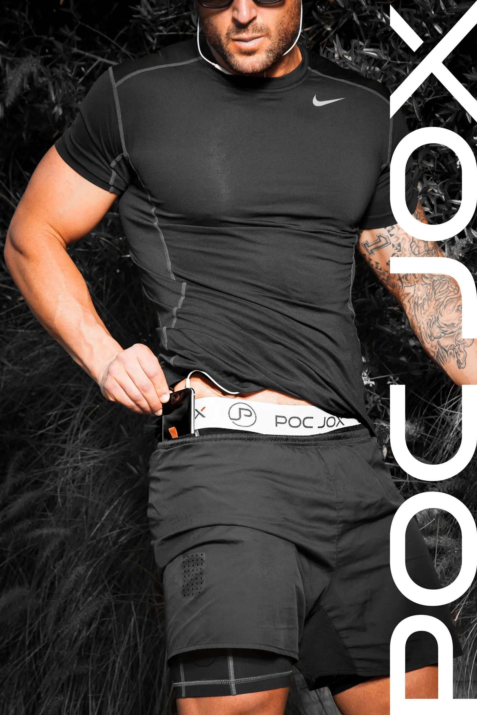 Man with muscular build in black athletic wear pulling the waistband of his shorts, showing the “POCJOX” branded underwear. He is also wearing earphones.