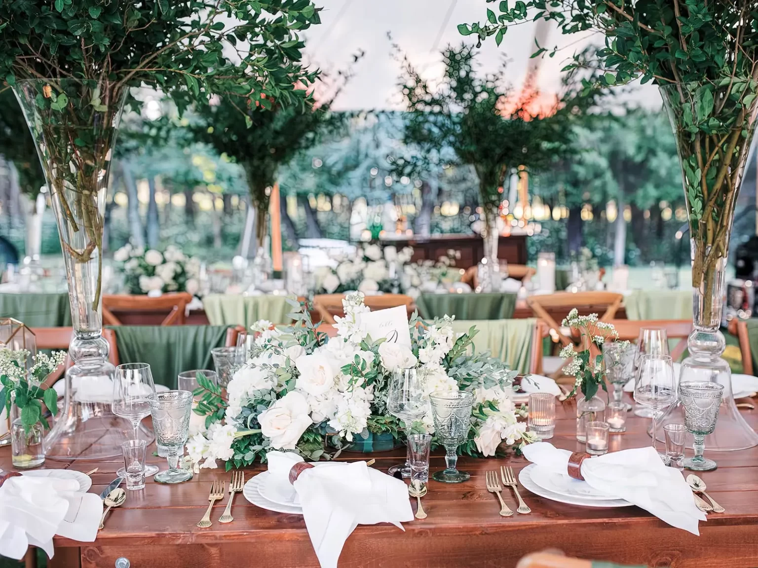 A beautifully set wooden table with white flowers, green foliage centerpieces, and green tablecloths under a tent, ready for an event.