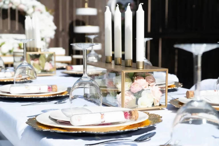An elegant table setting with white plates, gold chargers, folded napkins, inverted wine glasses, and a floral centerpiece with tall white candles.