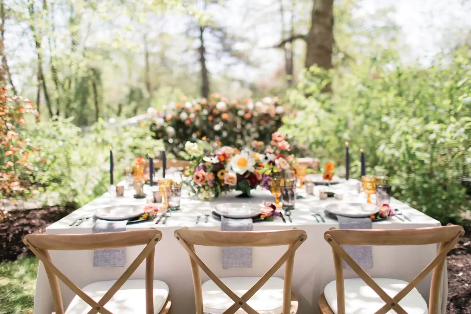 A white tablecloth with place settings and floral arrangements set for an outdoor event, surrounded by greenery and trees. Three wooden chairs are in the foreground.