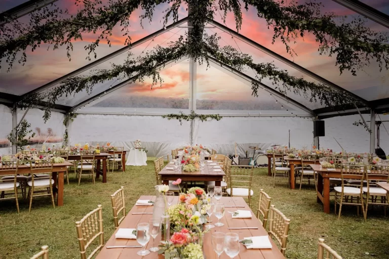 A wedding reception under a clear marquee with long wooden tables decorated with flowers and greenery, gold chairs, and a sunset sky visible through the tent roof.