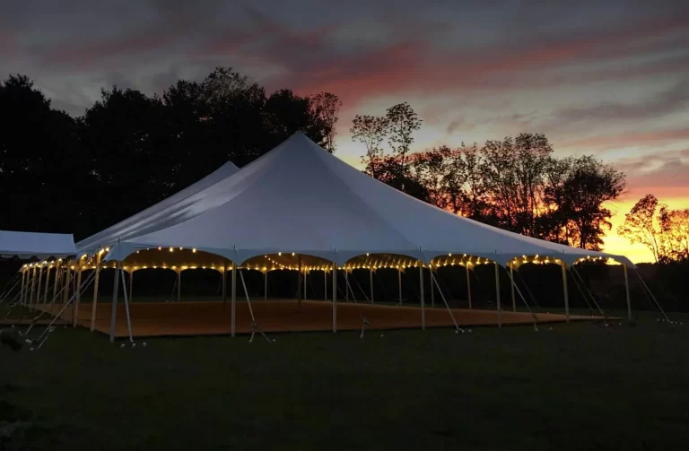 A large white tent with string lights is set up outdoors at dusk, surrounded by trees and under a colorful sky.