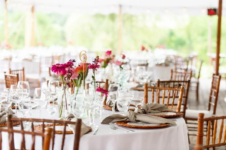 A banquet hall set with round tables adorned with pink floral centerpieces, white tablecloths, and wooden chairs. Numbered table cards and place settings with glassware and napkins are neatly arranged.