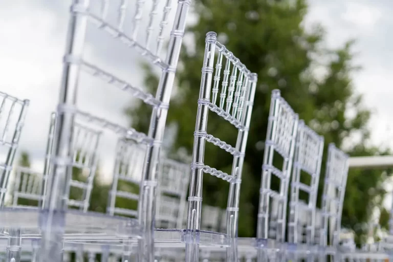 Rows of transparent Chiavari chairs are set up outdoors with greenery in the background.
