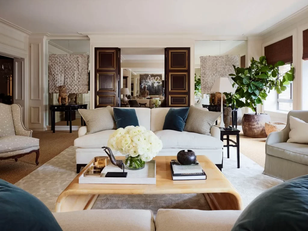 An elegant living room with a white sofa, wooden coffee table, plush chairs, and plants. Decor includes a vase of white flowers and books, with large mirrors and windows adding light to the space.