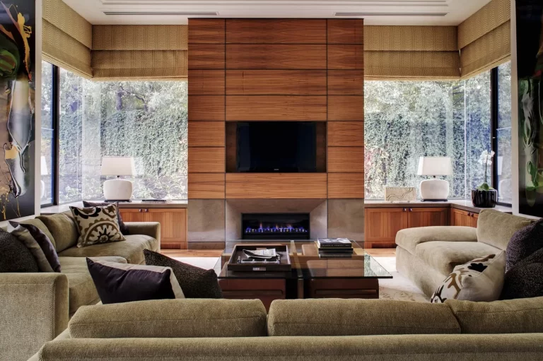 A modern living room featuring a wall-mounted TV above a fireplace, surrounded by large windows with shades, two sofas, and a coffee table in the center.