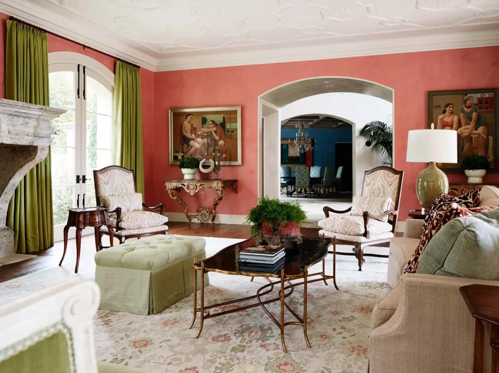 A well-furnished living room with coral walls, green curtains, a fireplace, two armchairs, a glass coffee table, framed artwork, and a view into a dining room.