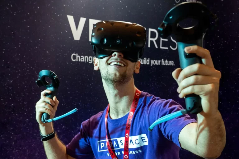 Person wearing a virtual reality headset and holding controllers, smiling with an open mouth, in front of a backdrop that includes the words "VR" and "build your future.