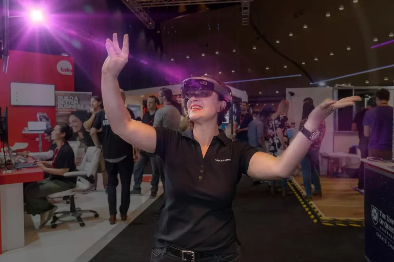 A person wearing a virtual reality headset interacts with a holographic display at a technology expo, surrounded by other attendees and various booths.