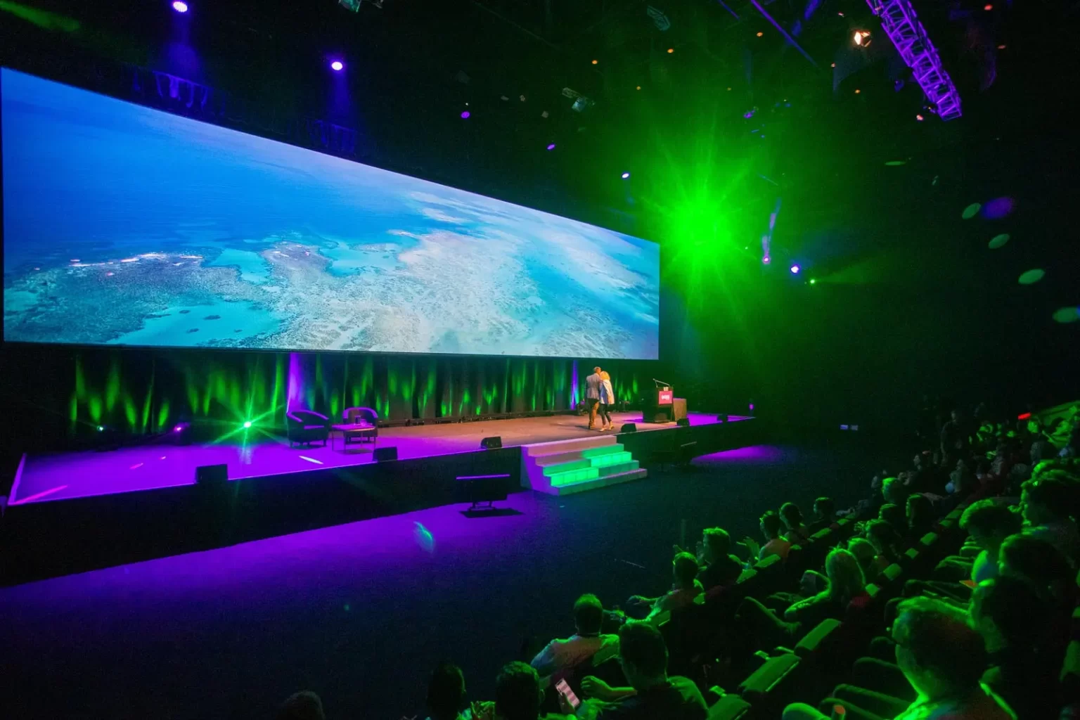 A man presents on a large stage with a panoramic screen displaying an aerial view of a coastal landscape, under green and purple stage lighting, to an audience in a darkened room.