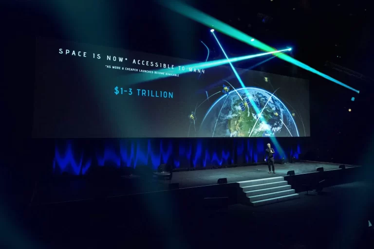 A speaker stands on a stage in front of a large screen displaying a presentation titled "Space is Now Accessible to Many" with projected economic figures of $1-3 trillion.