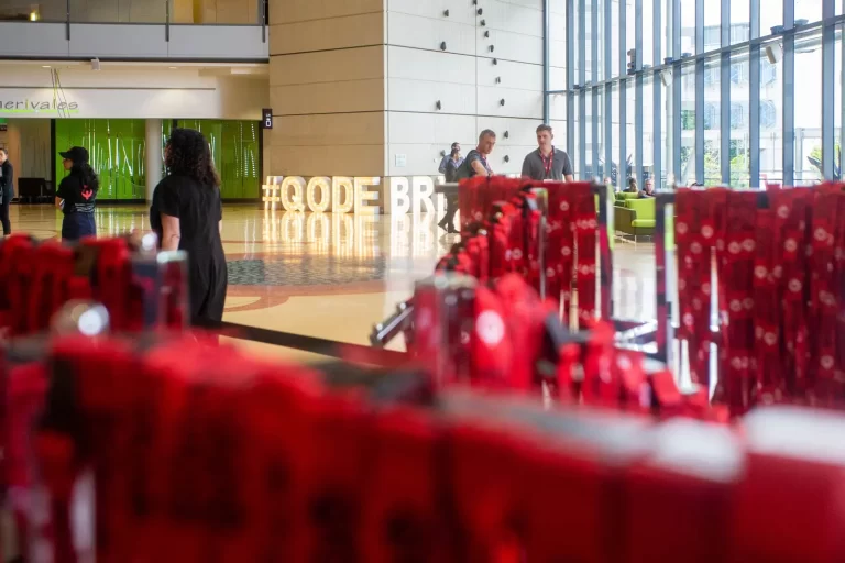 People interacting in a large, sunlit lobby with a "#CODE" sign in the background and red lanyards displayed in the foreground.