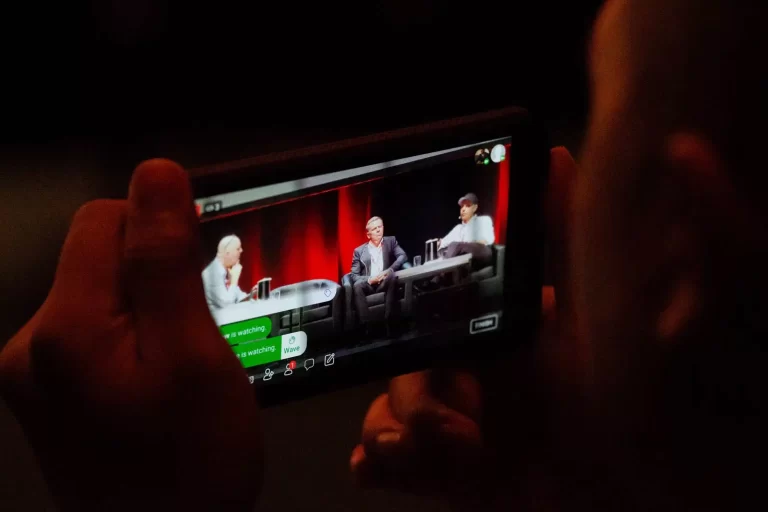 Person holds a phone showing a live stream of three individuals seated on a stage, engaged in a discussion with red curtains in the background.