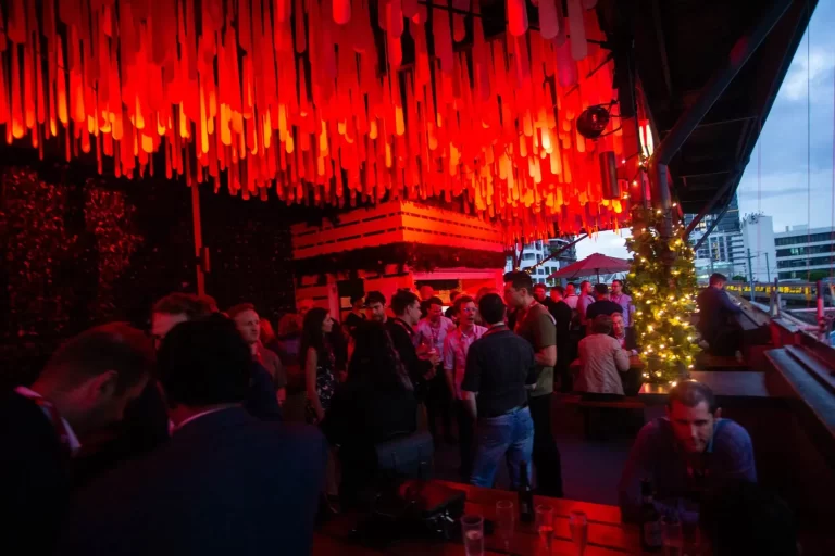 A group of people socializing at an outdoor evening event under hanging red lights, near a decorated tree and seating area.