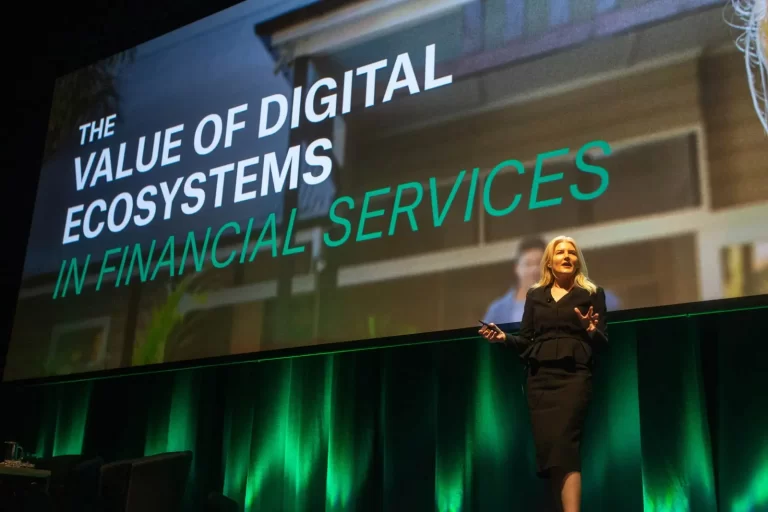 A speaker presents on stage with a large screen behind reading "The Value of Digital Ecosystems in Financial Services.