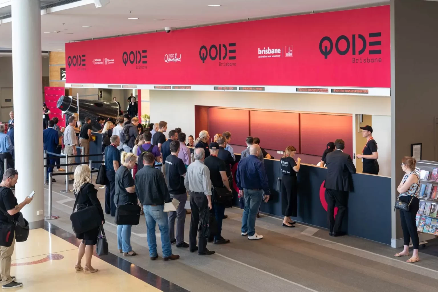 A crowd of people stands in line at a registration desk under a large red banner that reads "QODE Brisbane" in a convention center.