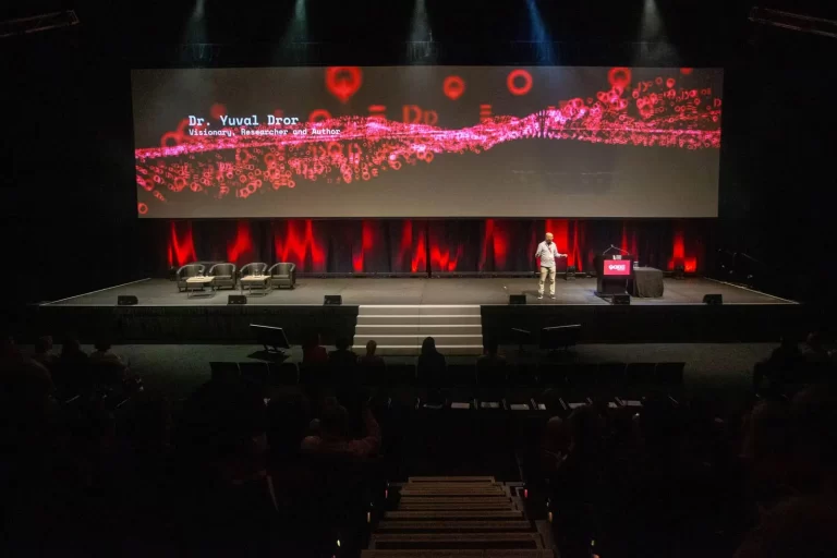 A speaker stands on a stage with a screen displaying the name "Dr. Yuval Dror" and a red, abstract pattern behind them. Several empty chairs are on stage, and audience seating is visible in the foreground.