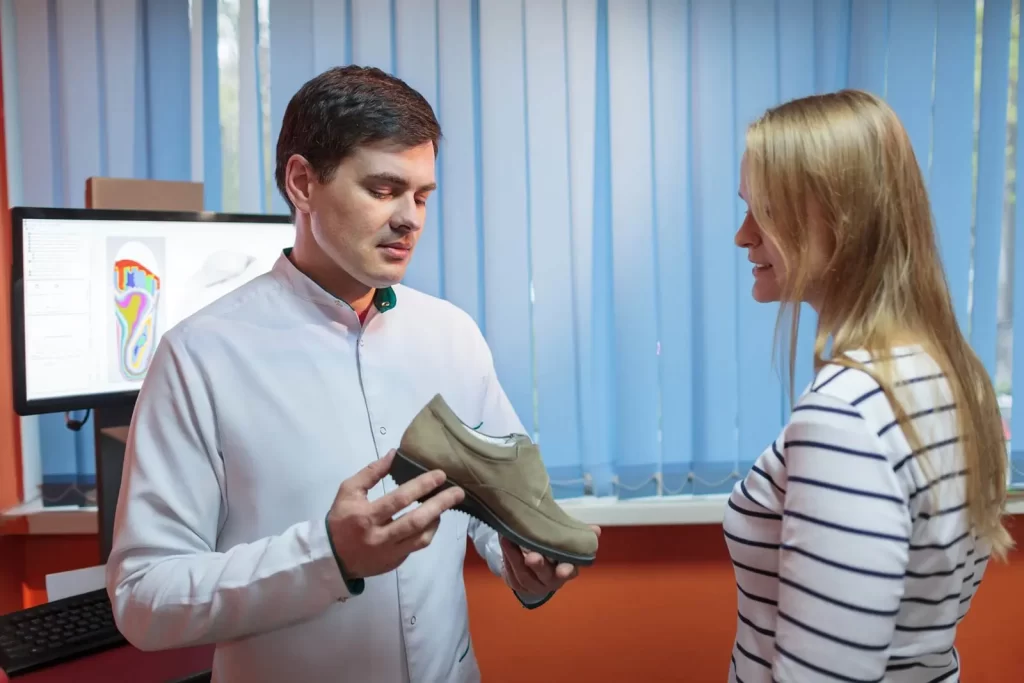 A man in a lab coat shows a shoe to a woman in a striped shirt, with a computer displaying a foot scan in the background.