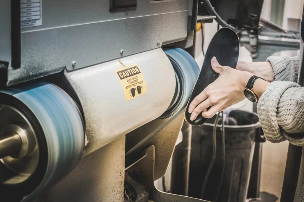 A person operates a shoe polishing machine, holding a shoe sole close to the rotating brushes. A caution sign is visible on the machine.