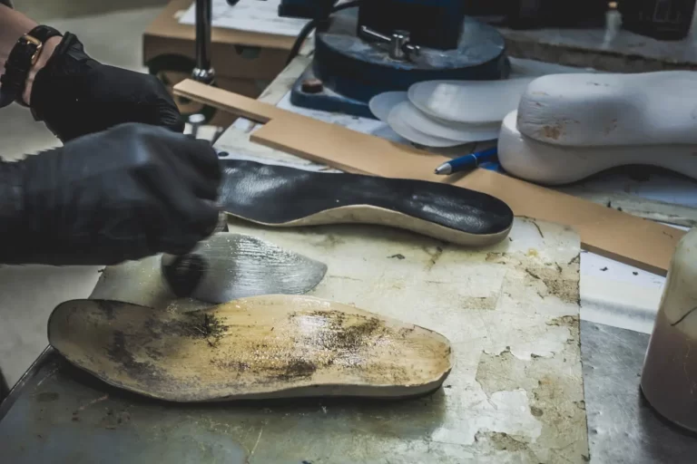 Person wearing black gloves applying adhesive to shoe insoles on a workbench, with shoe soles and tools in the background.