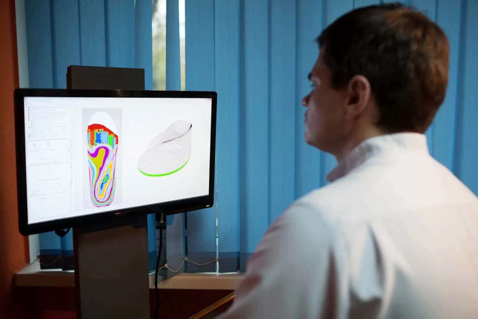 A person in a white coat looks at a computer screen displaying a 3D model and a colored contour map of a shoe.