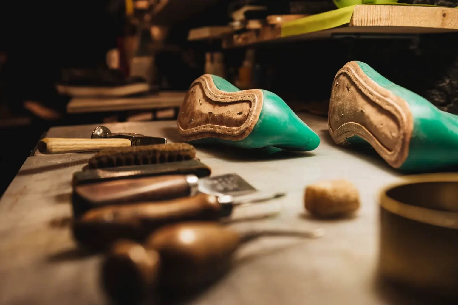 A cobbler's workspace with two green shoe lasts, a hammer, a brush, and other various leatherworking tools on a table.