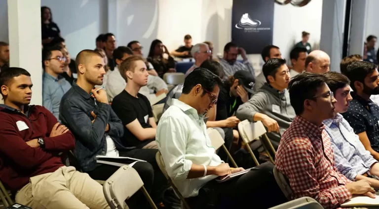 A group of people sitting in rows of chairs, some taking notes and others listening attentively, during a presentation or seminar in a well-lit room.