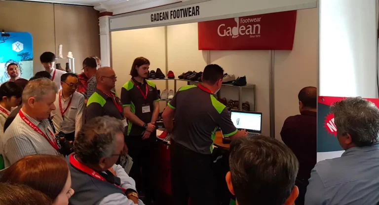 A group of people, including two demonstrators in gray and green shirts, gathers around a booth labeled "GADEAN FOOTWEAR" at an exhibition. One demonstrator shows something on a laptop screen.