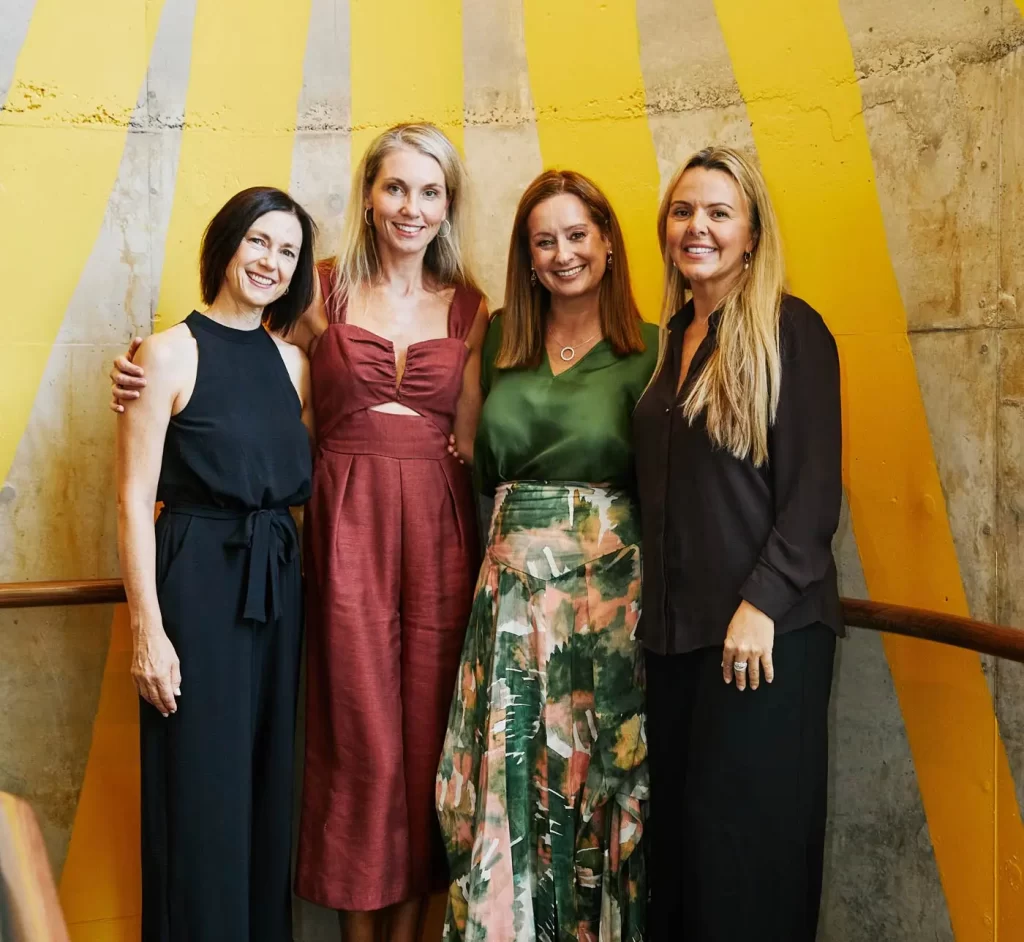 Four women stand together, smiling, in front of a yellow-striped wall. They are dressed in various styles, including dresses and a blouse with a skirt.