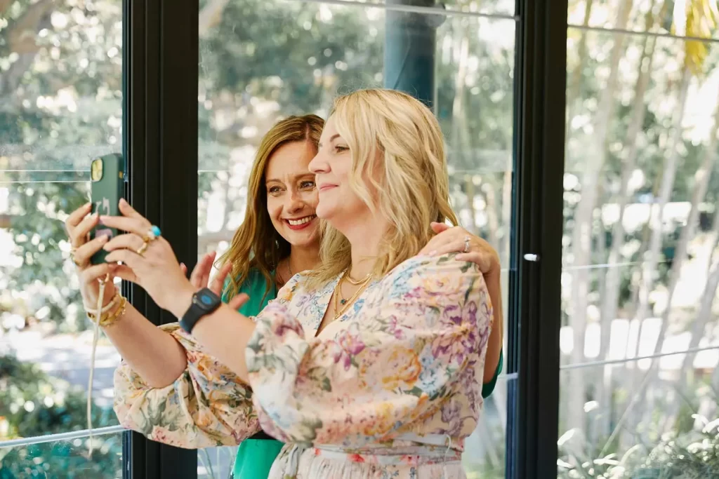 Two women are taking a selfie in front of a window. One woman is holding the phone, while the other stands beside her, smiling. Trees and sunlight are visible outside.