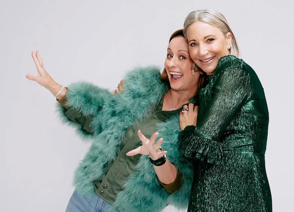 Two women pose playfully. One wears a green faux fur coat and jeans, and the other wears a shiny green dress. Both smile and appear cheerful against a plain background.