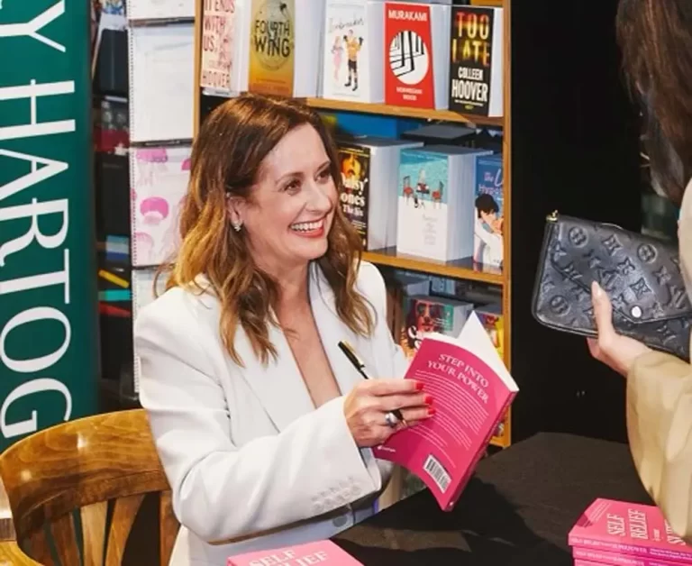 A woman in a white blazer smiles while signing a book at a book signing event. She is seated at a table with multiple copies of her book. Bookshelves are visible in the background.