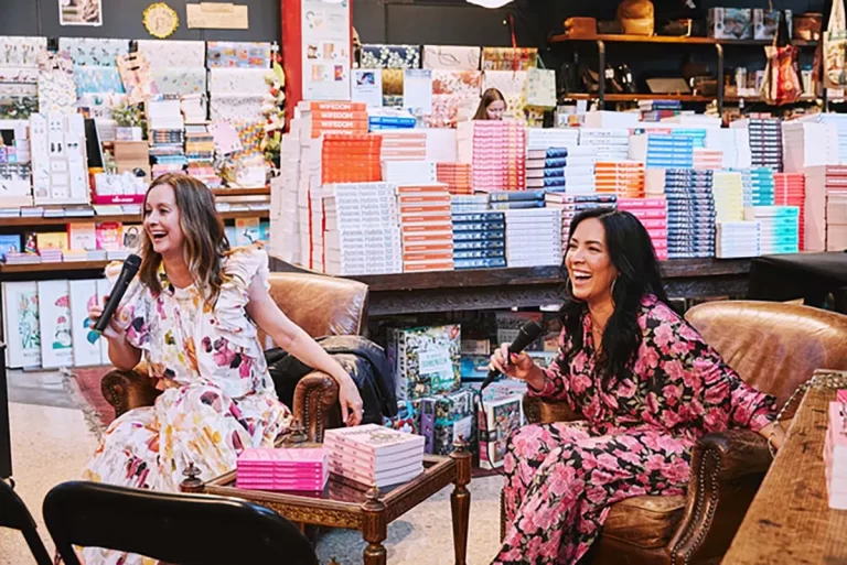 Two women in floral dresses are sitting and holding microphones in a bookstore with stacks of colorful books on the table and shelves behind them. They appear to be engaged in a lively conversation.