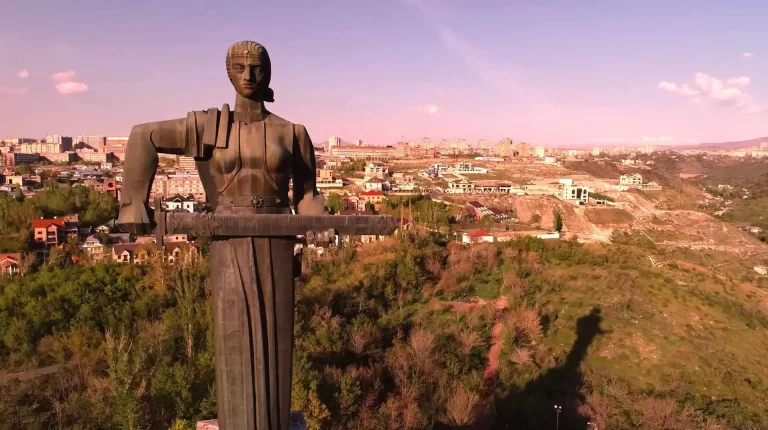 A large statue of a woman holding a sword and a cup stands prominently with a sprawling urban landscape and greenery visible in the background.